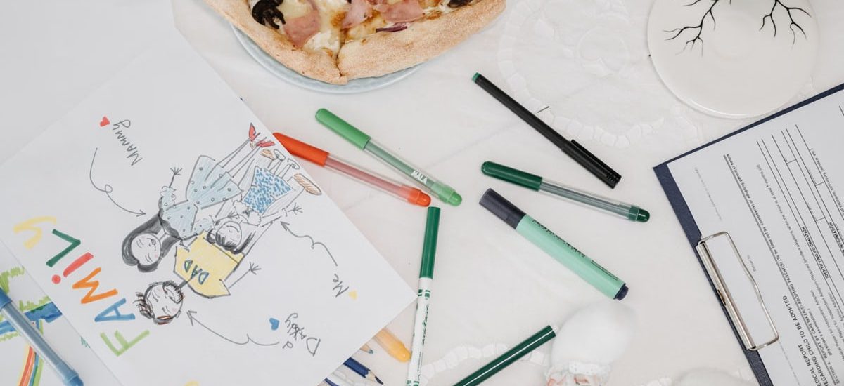 A colorful child's drawing of a family alongside pizza and pens on a table.